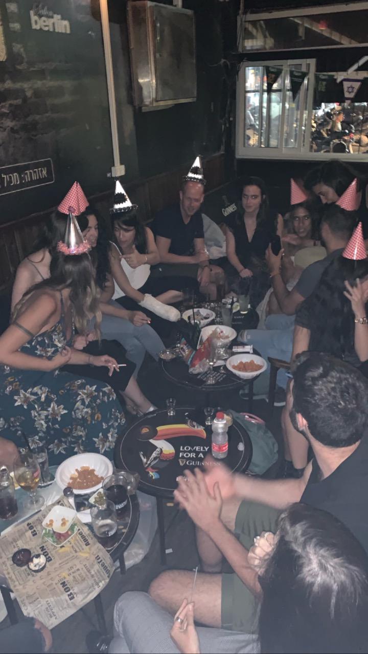 חבורת חברים עם כובעי יום הולדת סביב שולחנות קטנים בבר חשוך בסלון ברלין