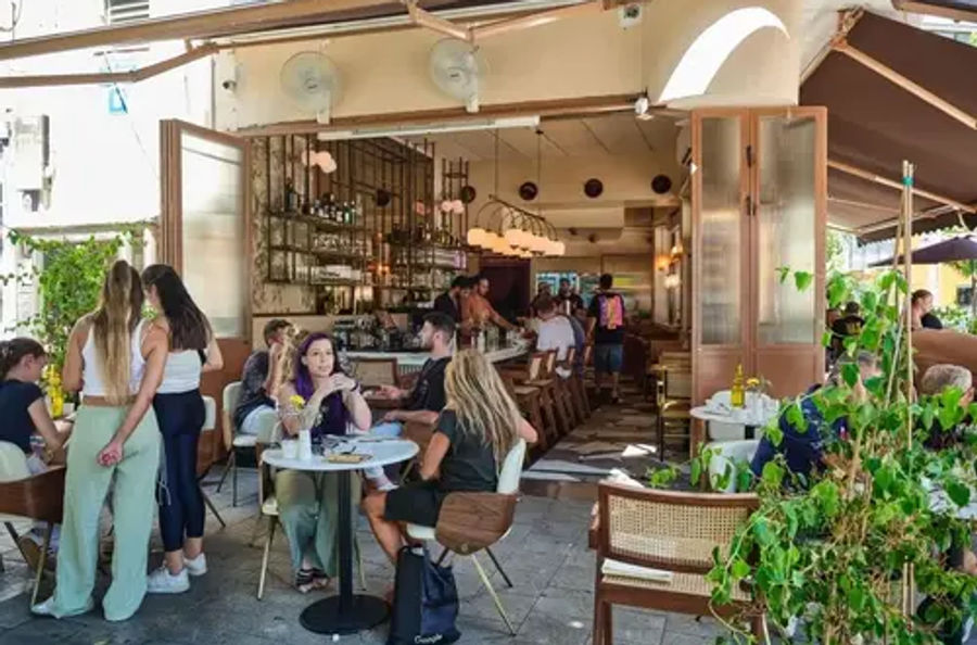 אנשים יושבים בשולחנות עגולים על המדרכה ובבר הפתוח של AT8, עם כיסאות קש ועציצים בכניסה